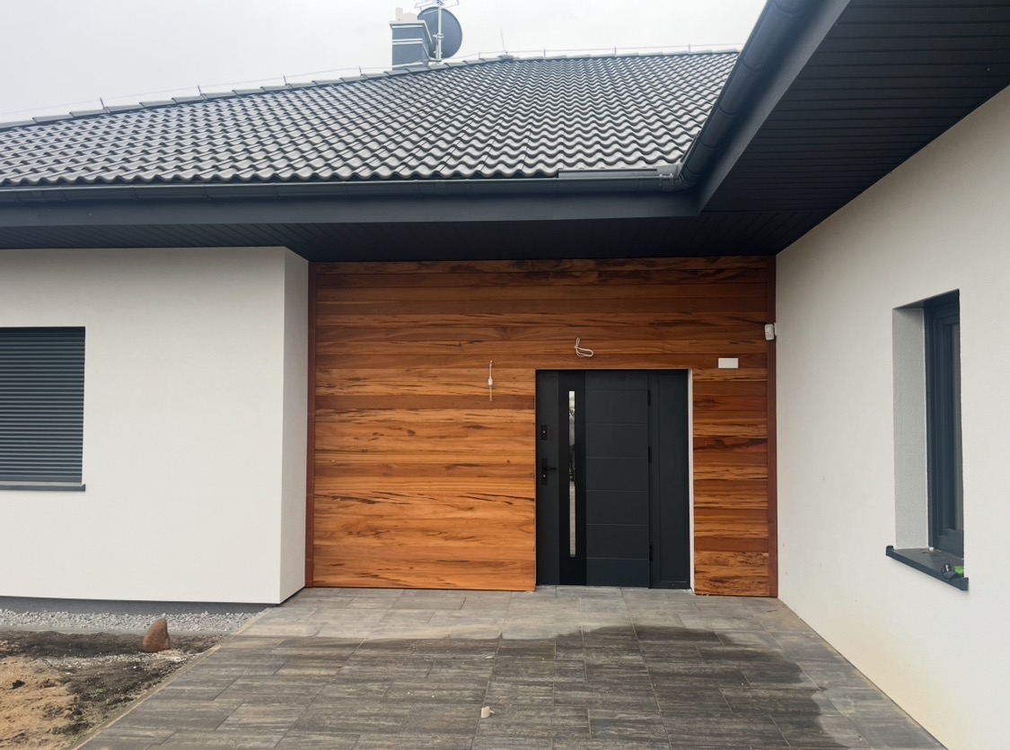 Nowoczesny dom jednorodzinny z elewacją wykończoną drewnem egzotycznym, antracytowe okna i drzwi, szara kostka brukowa na podjeździe, widoczne przewody elektryczne nad drzwiami.