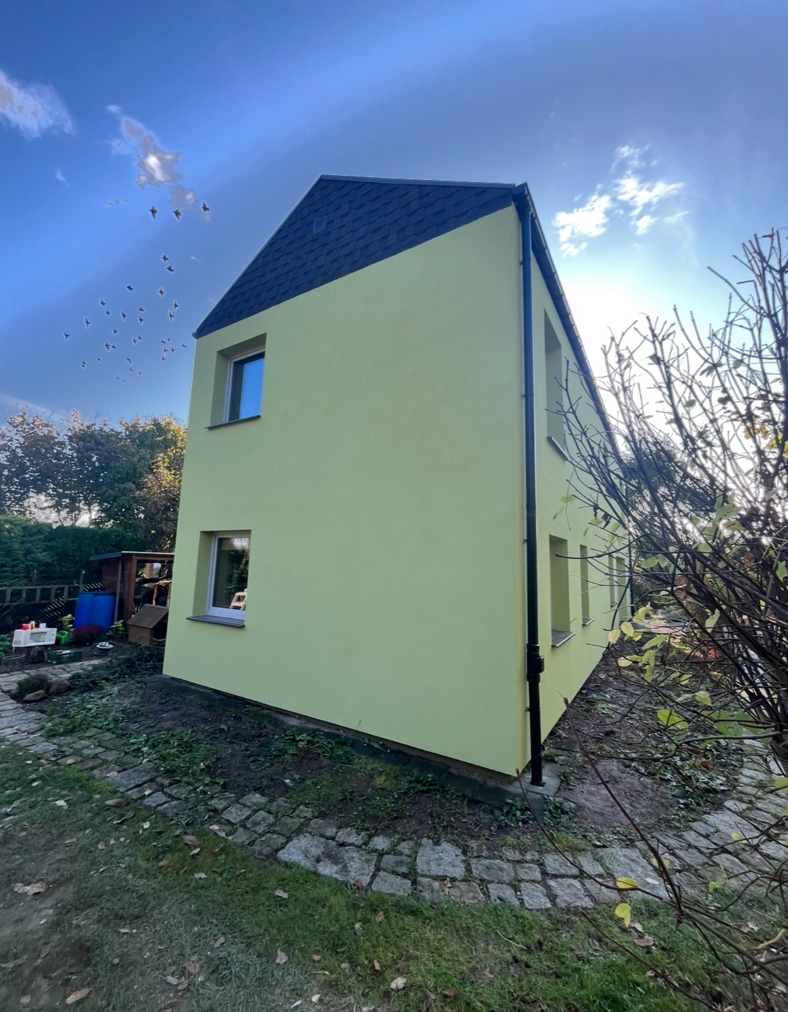 Dwupiętrowy dom z żółtą elewacją i czarnym dachem, widoczny z perspektywy ogrodu z kostką brukową i krzewami.