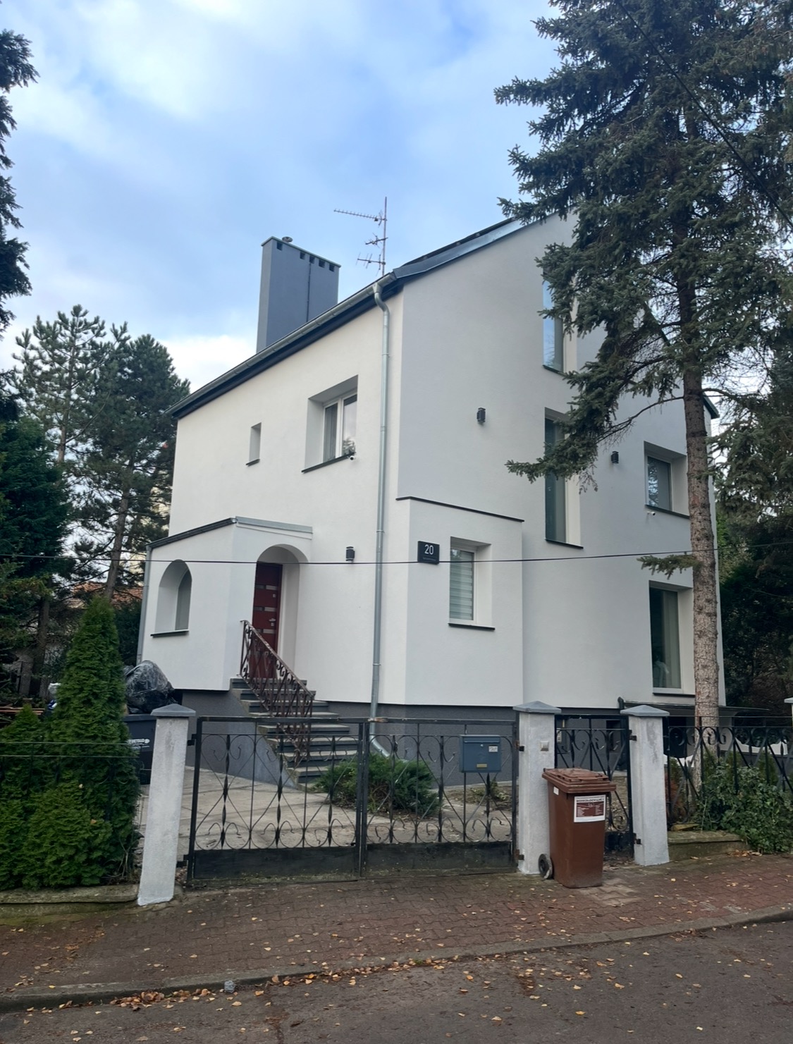 Dwupiętrowy dom z białą elewacją, ciemną stolarką okienną i numerem 20 na ścianie, widoczny zza metalowej bramy i ogrodzenia.
