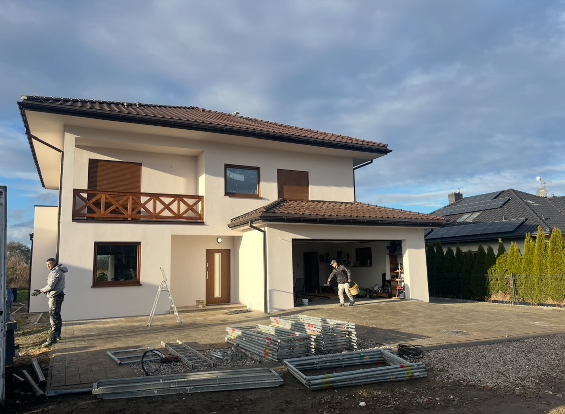 Dwukondygnacyjny dom jednorodzinny z białymi ścianami elewacyjnymi, brązowymi oknami i balkonem z drewnianą balustradą; na podjeździe leżą elementy rusztowania, a w otwartym garażu widać pracownika...