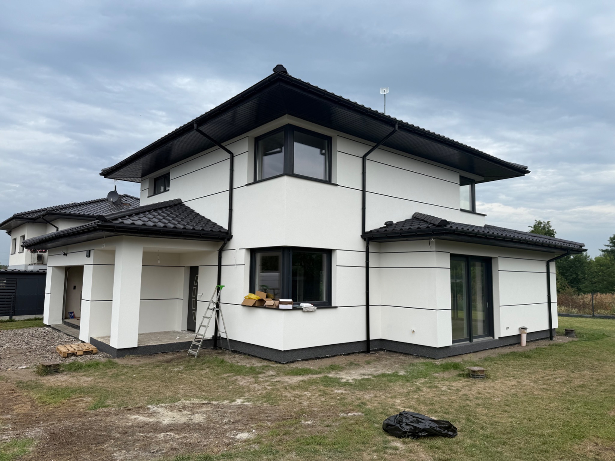 Nowoczesny dom z białą elewacją i czarnymi akcentami, widoczna drabina i materiały budowlane, sugerujące prace wykończeniowe. Pochmurne niebo w tle.