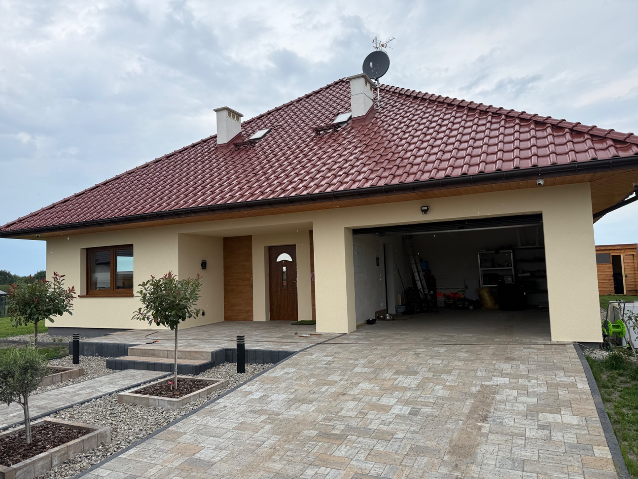 Elewacja domu jednorodzinnego w beżowym kolorze z czerwoną dachówką, widok na podjazd z kostki brukowej i otwarty garaż. Nowoczesny styl.