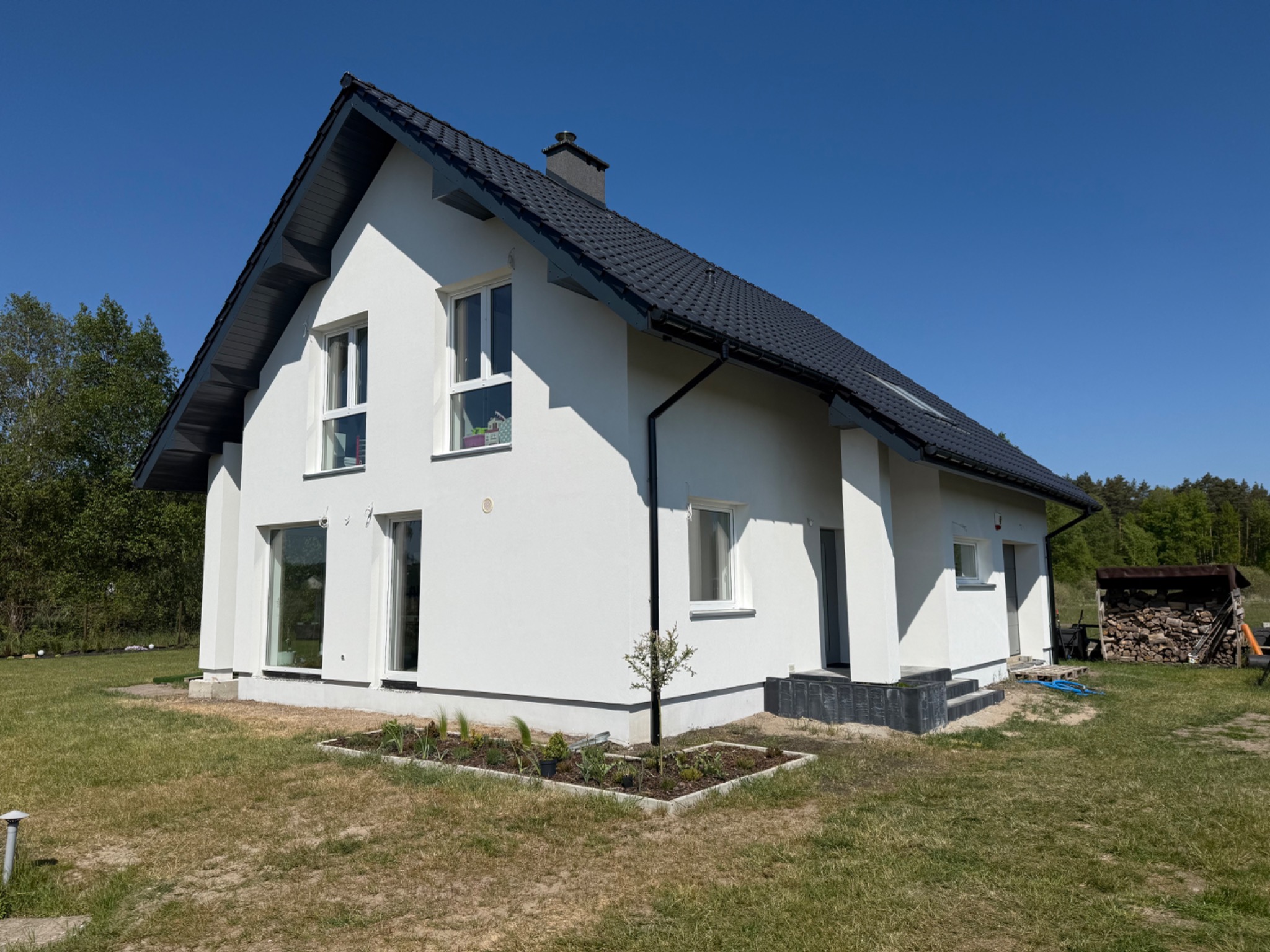 Nowoczesny dom jednorodzinny z białą elewacją i ciemną dachówką, widok z ogrodu w słoneczny dzień. Mały ogródek przed domem.