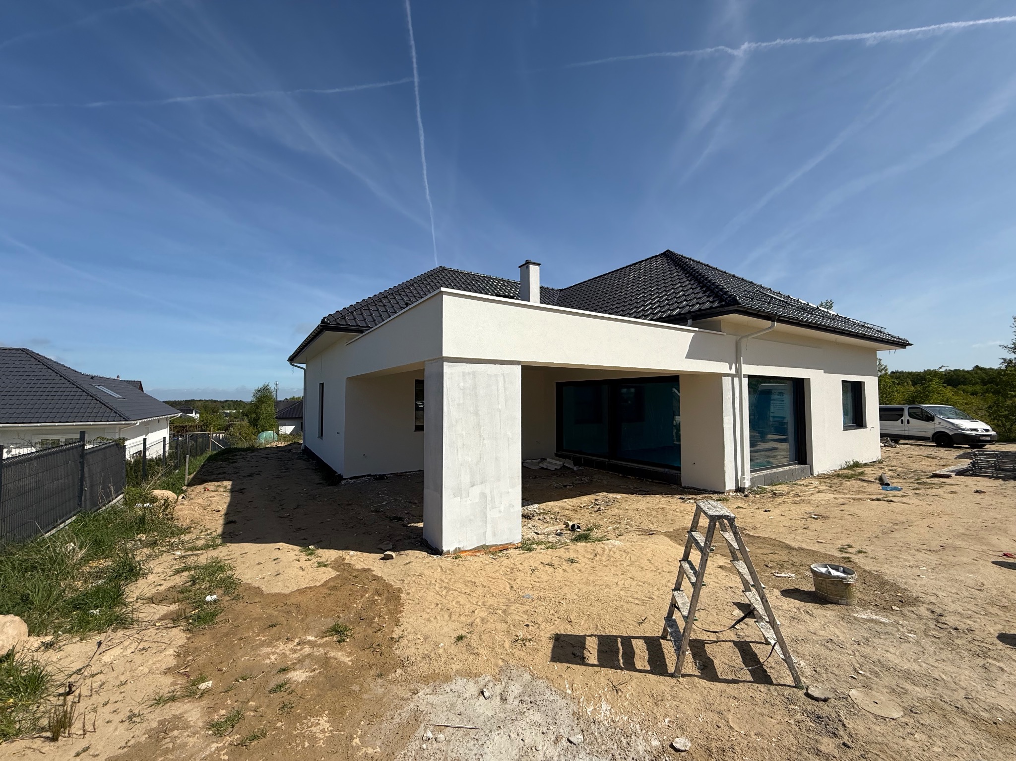 Nowoczesny, parterowy dom z białymi ścianami i czarnym dachem, widoczny z boku. Na pierwszym planie drabina i teren w trakcie prac wykończeniowych.