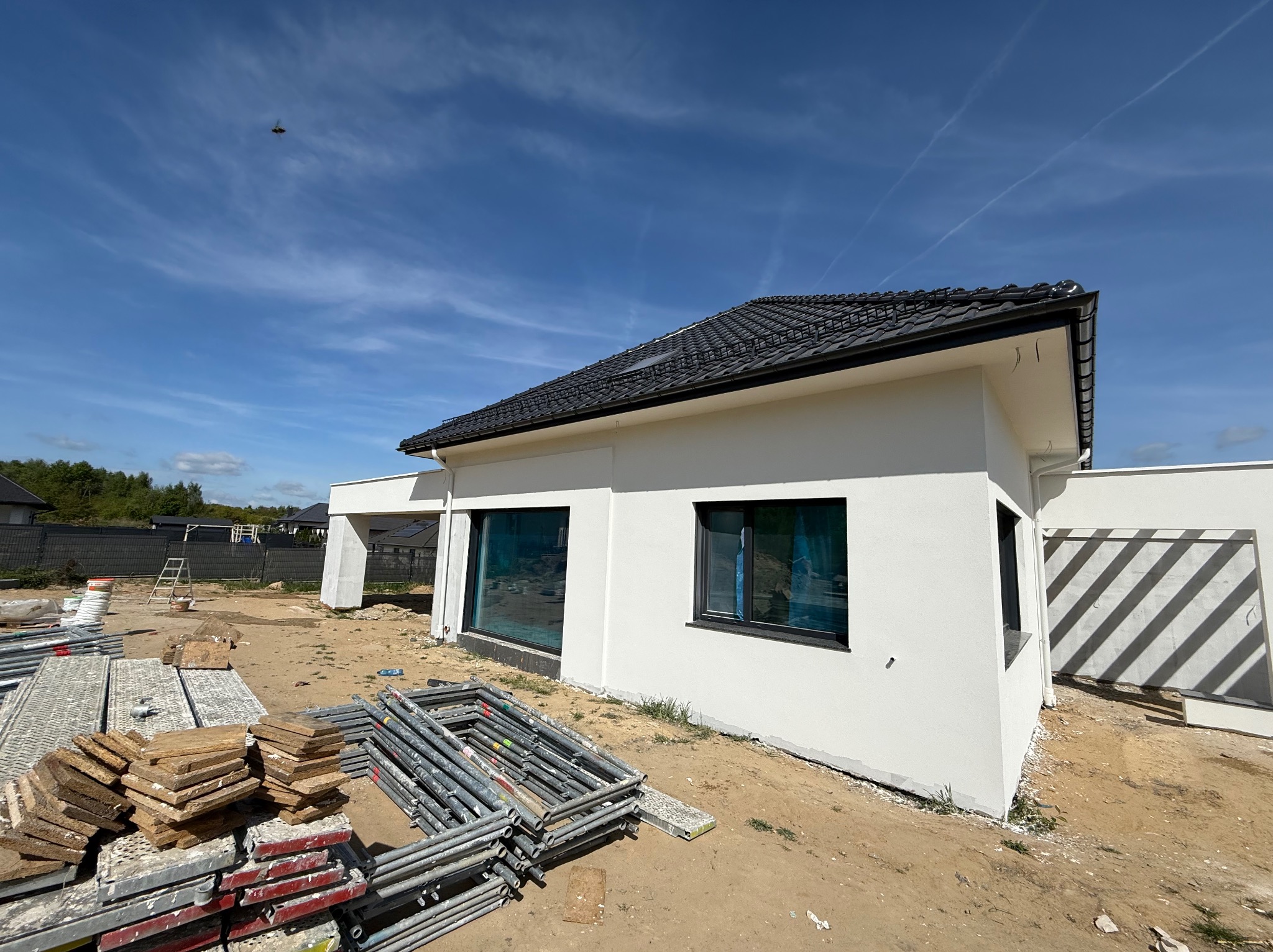 Nowoczesny, parterowy dom z białymi ścianami i czarną dachówką w trakcie prac wykończeniowych. Widoczne rusztowania i materiały budowlane na posesji.