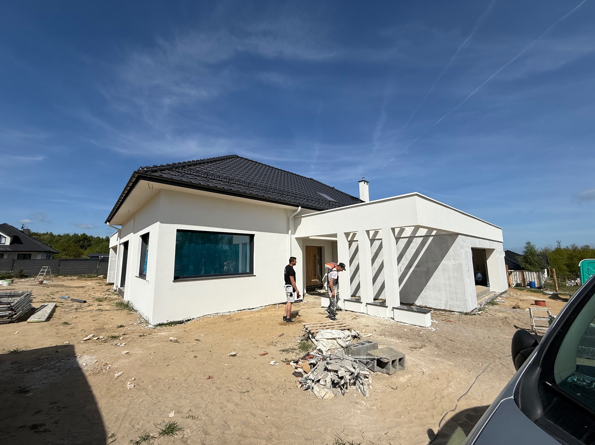 Biały dom jednorodzinny z czarnym dachem w trakcie prac wykończeniowych elewacji. Widoczni dwaj pracownicy i gruz budowlany na posesji.