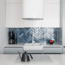 SUSPENS studio - Nowoczesna kuchnia z niebieskimi płytkami ułożonymi w jodełkę, białymi szafkami i blatem. Na blacie czajnik i ekspres do kawy. Obok stoi abstrakcyjna rzeźba.