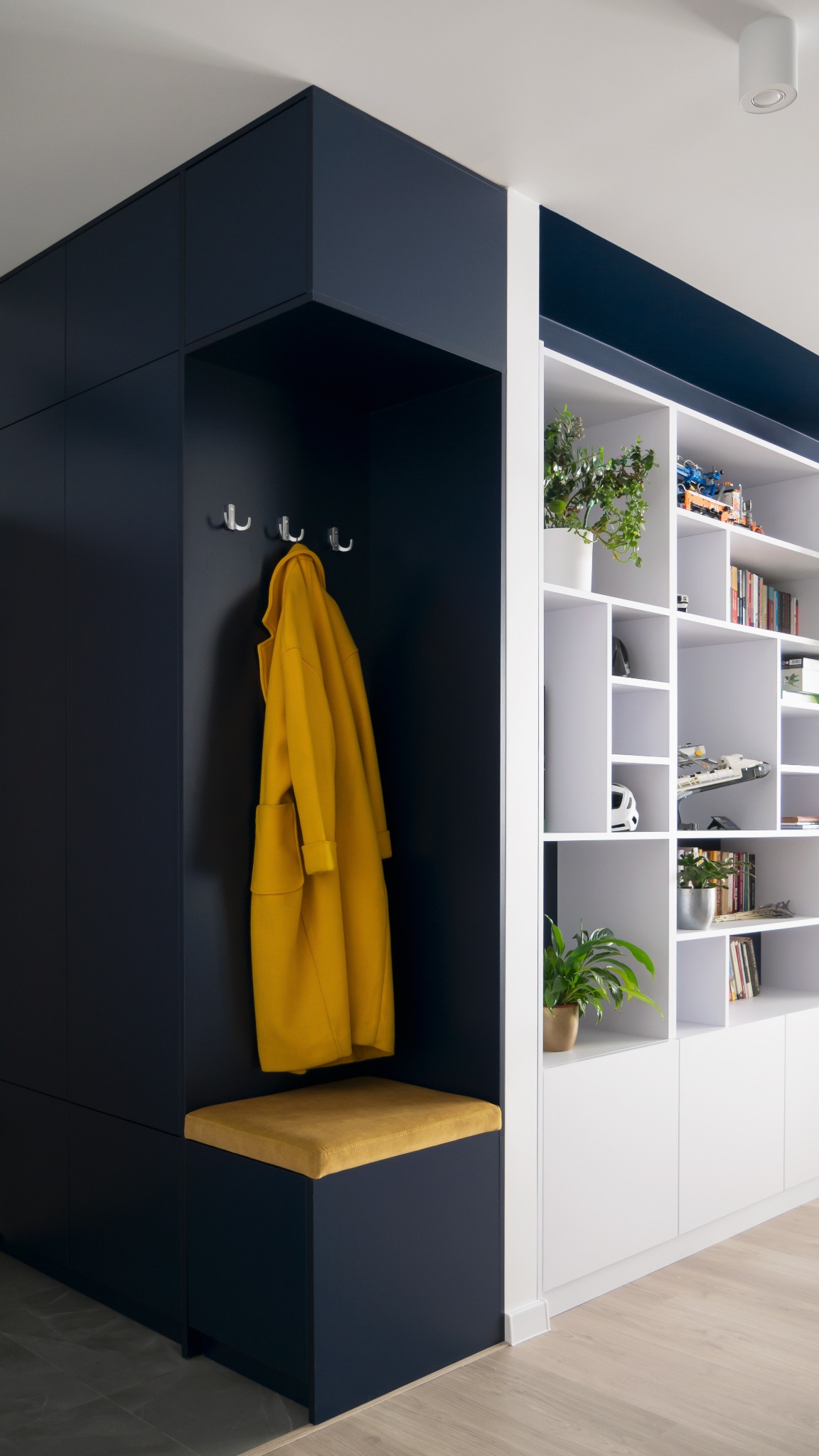 Nowoczesna zabudowa przedpokoju: granatowe szafy z siedziskiem i wieszakami, biały regał z roślinami i dekoracjami. Żółty płaszcz dodaje koloru.