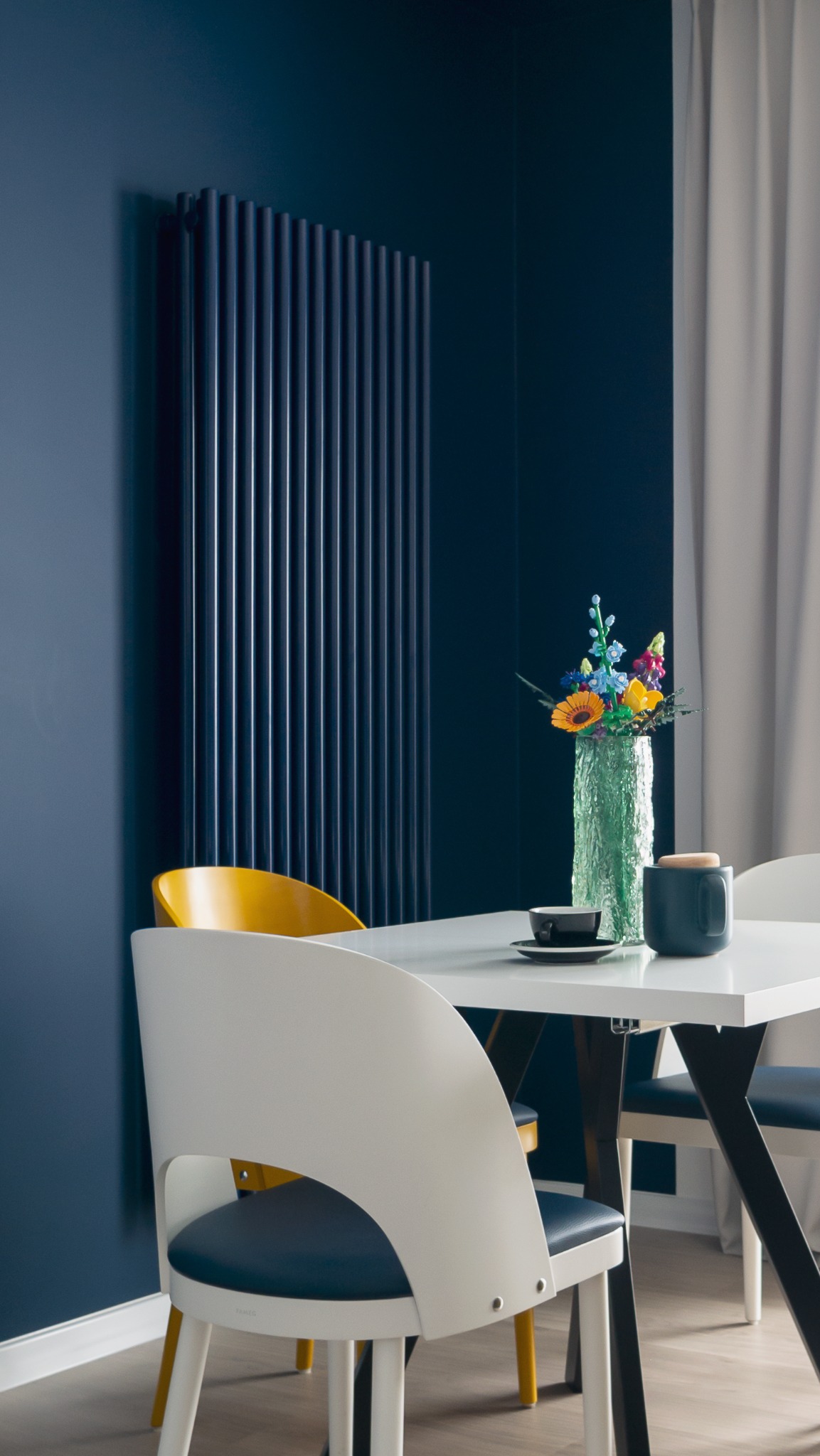 Nowoczesne wnętrze: granatowa ściana z grzejnikiem, biały stół z kwiatami, biało-żółte krzesła i szare zasłony. Minimalistyczny design.