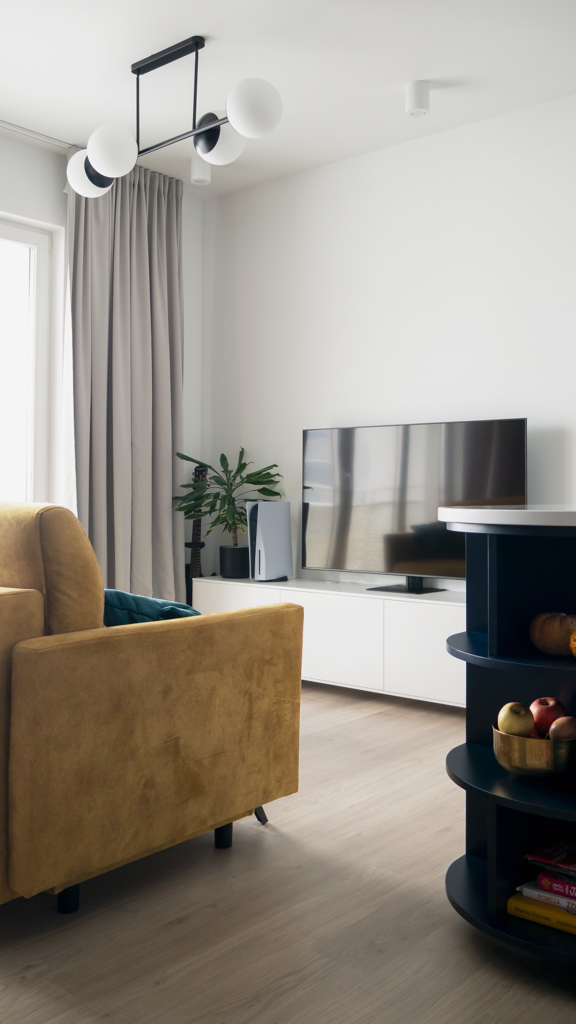 Nowoczesny salon z musztardową sofą, białą szafką RTV, konsolą PS5 i okrągłym regałem w odcieniach granatu. Minimalistyczna lampa sufitowa.