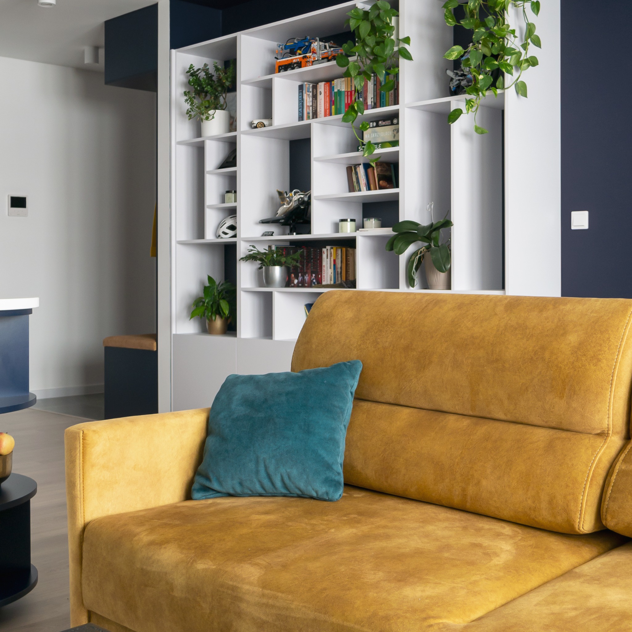 Nowoczesny salon z żółtą sofą i turkusową poduszką. W tle biały regał z roślinami i książkami, granatowa ściana. Widoczny fragment wnętrza mieszkania.