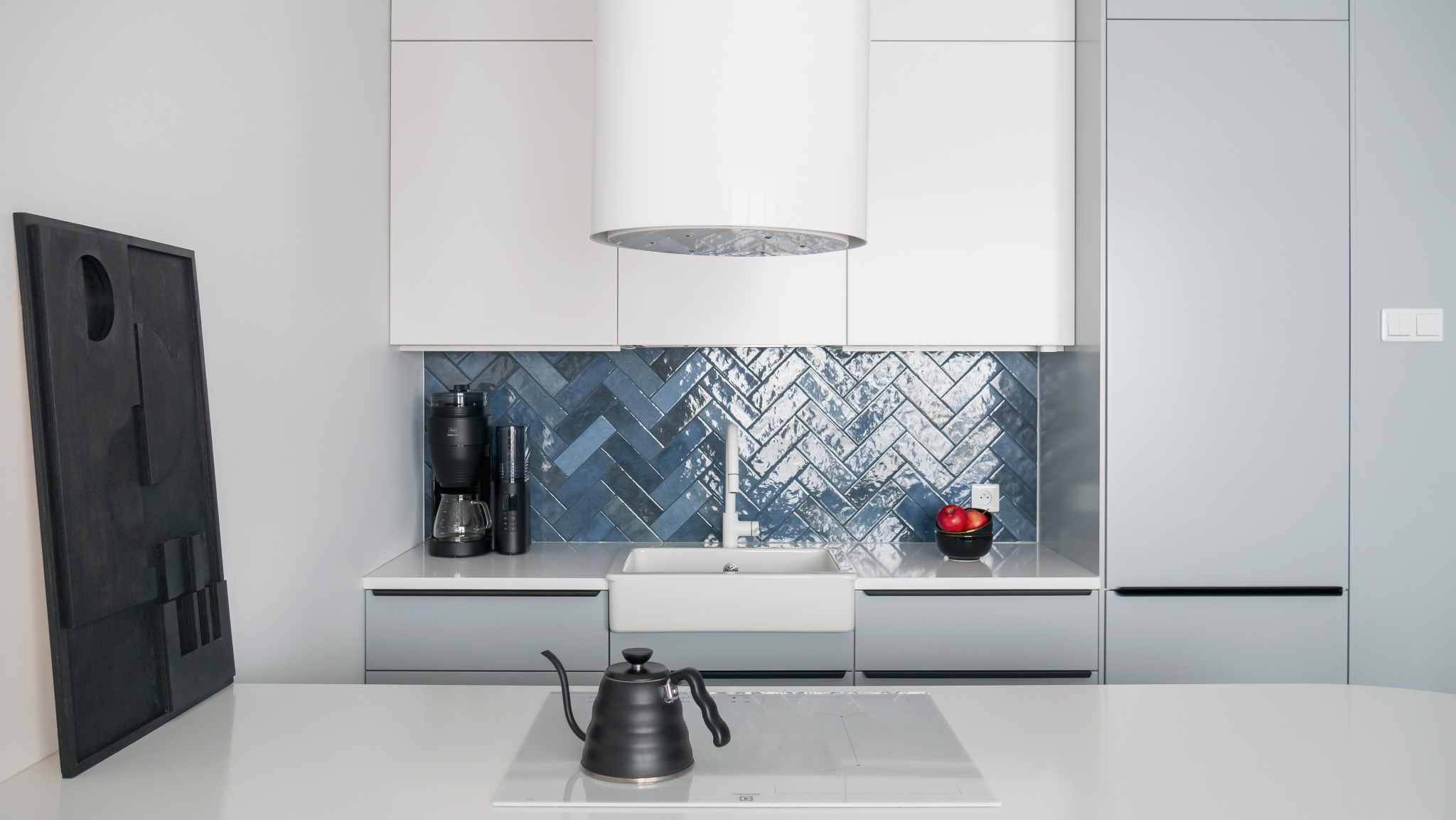 Nowoczesna kuchnia z niebieskimi płytkami ułożonymi w jodełkę, białymi szafkami i blatem. Na blacie czajnik i ekspres do kawy. Obok stoi abstrakcyjna rzeźba.