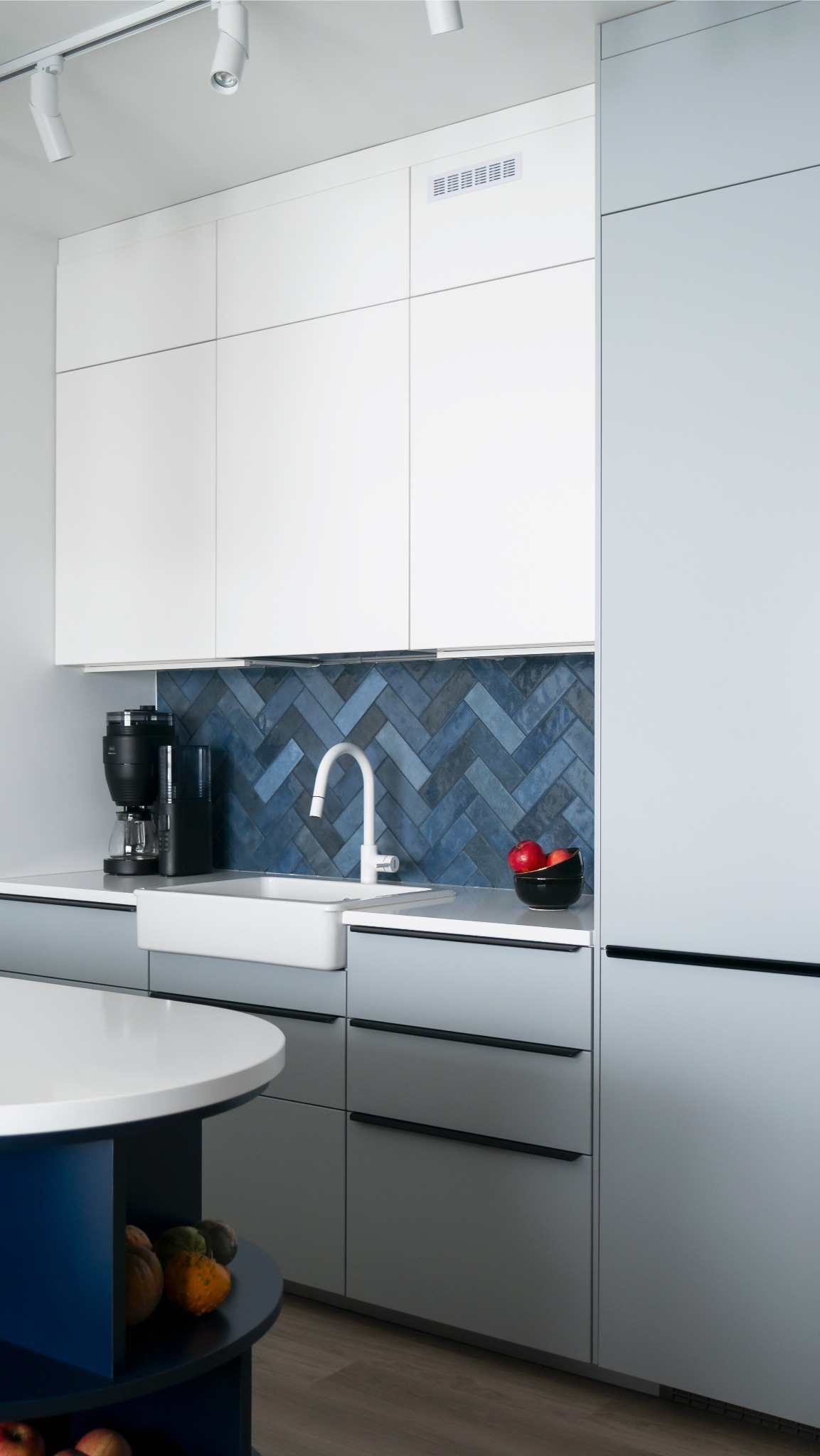 Nowoczesna kuchnia: białe szafki, niebieska mozaika w jodełkę nad zlewem, czarny ekspres do kawy i owocowa misa. Minimalistyczny design, jasne oświetlenie.