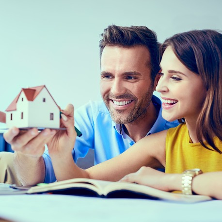 Uśmiechnięta para trzyma w dłoniach model domu z czerwoną dachówką, przeglądając otwartą książkę, prawdopodobnie planując przyszłe inwestycje.