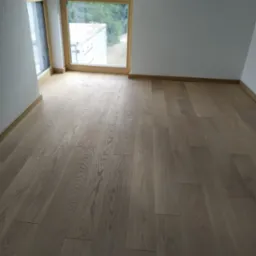 Jasne, drewniane panele podłogowe ułożone w nowym, pustym pokoju z dużym oknem, widok na zieleń na zewnątrz.