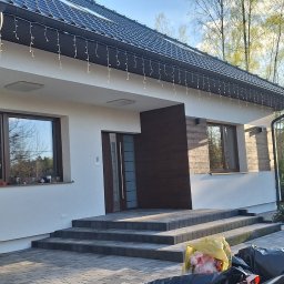 REM-SAR - Nowoczesny dom jednorodzinny z białą elewacją i elementami dekoracyjnymi imitującymi drewno, widoczne schody wejściowe wyłożone kostką brukową, w tle drzewa i elementy ogrodu.