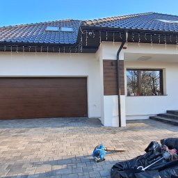 REM-SAR - Nowoczesny dom jednorodzinny z białą elewacją, brązową bramą garażową i drzwiami wejściowymi, czarnym dachem z oknami dachowymi, oświetleniem zewnętrznym, kostką brukową na podjeździe...