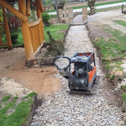 REM-SAR - Pomarańczowa zagęszczarka wibrująca ubija warstwę białego kruszywa na przygotowanej ścieżce ogrodowej, widoczna drewniana altana z boku i stos kruszywa w tle.