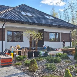 REM-SAR - Nowoczesny dom jednorodzinny z elementami drewnianymi na elewacji, widok na ogród z kostki brukowej i roślinności, pracownik w oddali przy elewacji, drugi pracownik idzie w stronę domu.