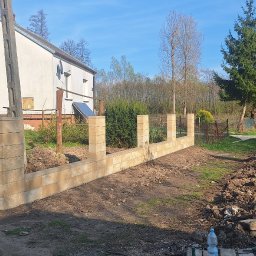 REM-SAR - Wznoszona z beżowych bloczków podmurówka ogrodzenia na tle wiejskiego domu z instalacją solarną i zielenią, widoczne narzędzia i butelka wody na palecie.