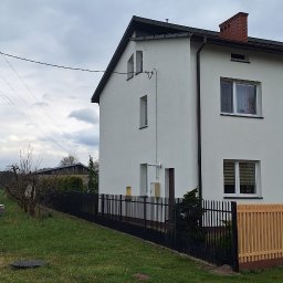 REM-SAR - Dwukondygnacyjny dom z białą elewacją, czarną dachówką i ceglanym kominem, otoczony czarnym metalowym ogrodzeniem z drewnianym panelem, widok z poziomu trawnika w pochmurny dzień.