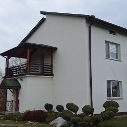 REM-SAR - Dwukondygnacyjny dom z białymi ścianami po ociepleniu, z drewnianym balkonem i ozdobnymi krzewami w ogrodzie, widoczne pozostałości materiałów budowlanych.