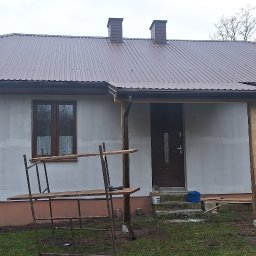 REM-SAR - Dom jednorodzinny w trakcie prac wykończeniowych elewacji, widoczne rusztowania, pracownicy na drabinie oraz fragment ocieplonego poddasza z brązowym dachem.