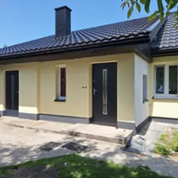 Dom jednorodzinny z nową elewacją w odcieniach beżu i szarości, czarny dach z dachówki ceramicznej, widoczny fragment pracownika przy elewacji bocznej.
