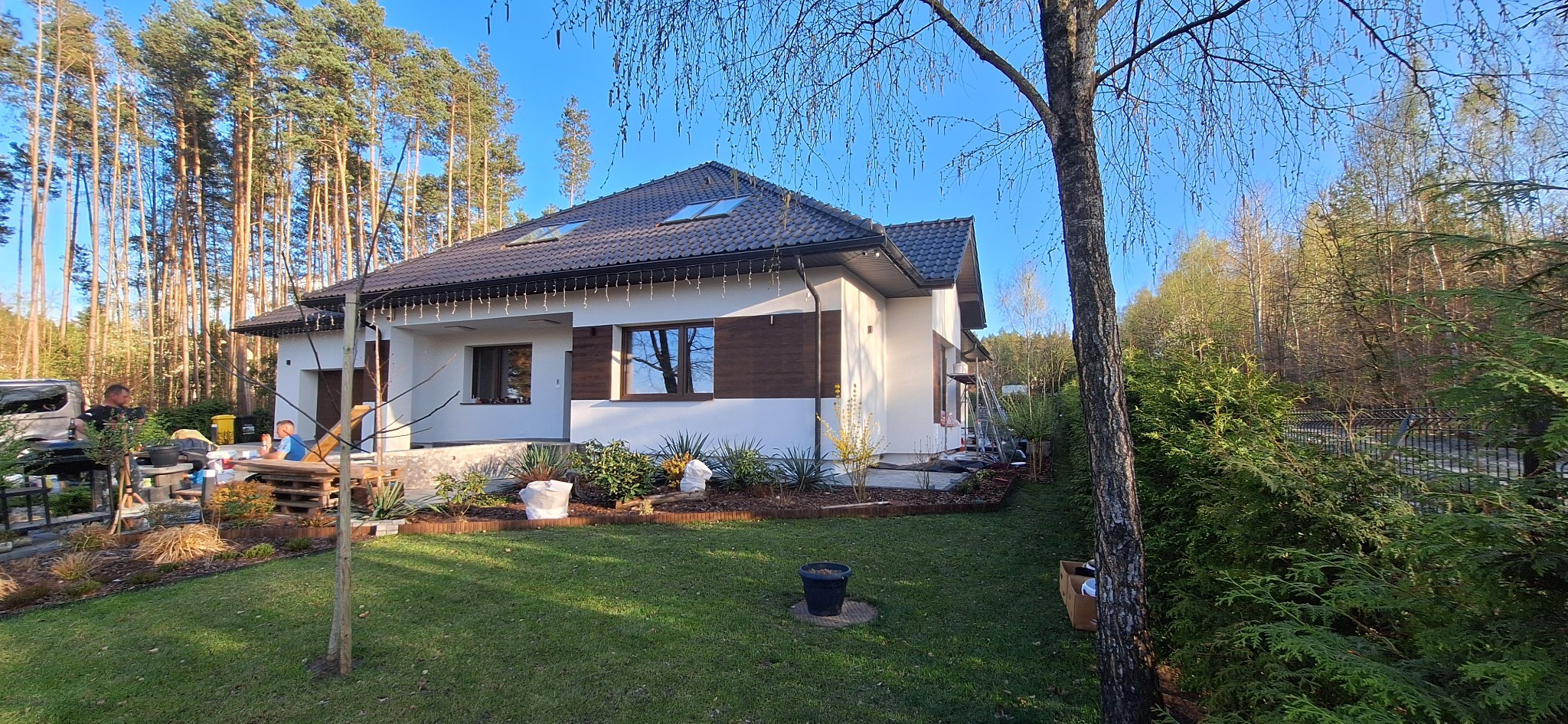 Nowoczesny dom jednorodzinny z elewacją w kolorze białym i brązowymi akcentami, widoczne okna z brązowymi ramami, ciemny dach z oknami dachowymi, teren wokół domu zagospodarowany zielenią...