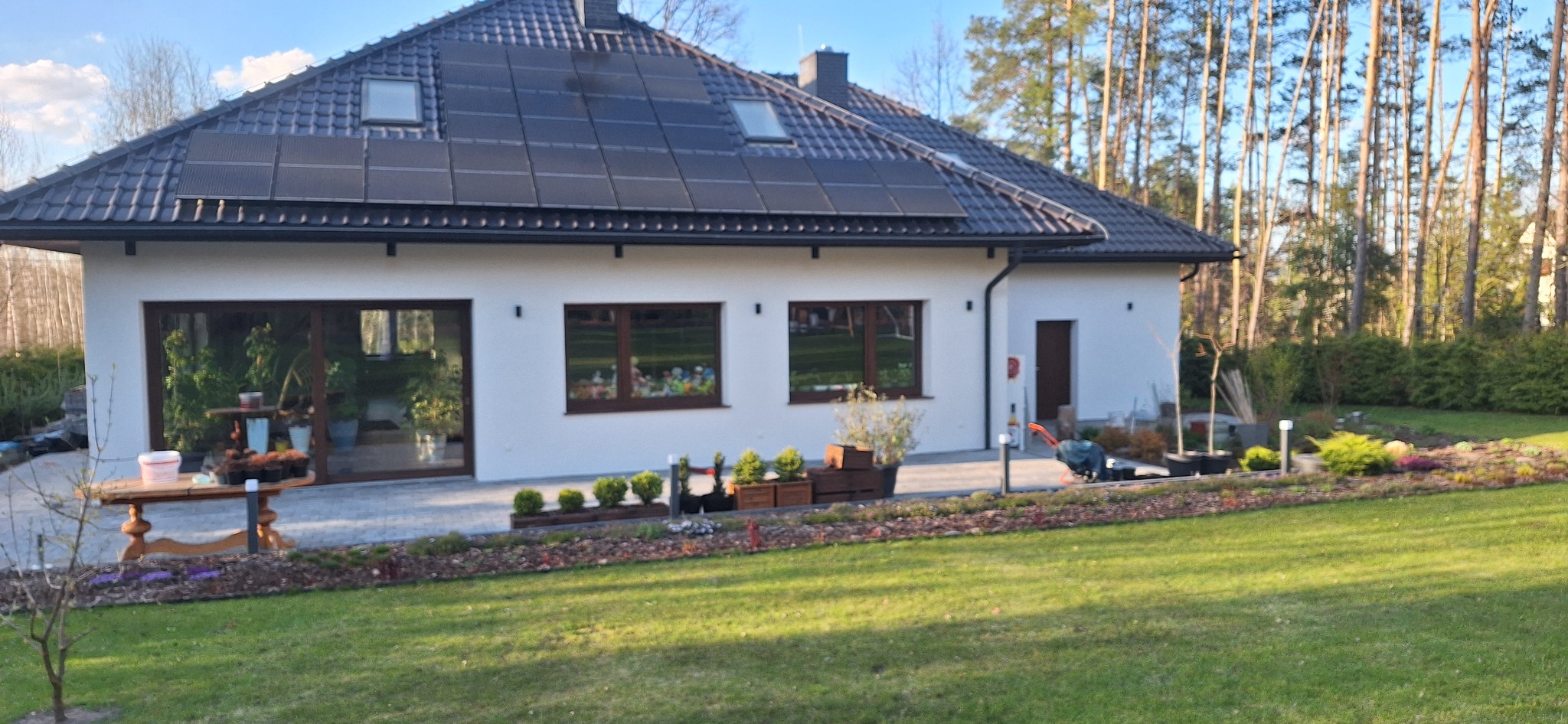 Nowoczesny dom jednorodzinny z białymi ścianami, brązowymi ramami okiennymi i panelami słonecznymi na dachu, otoczony zadbanym ogrodem z elementami dekoracyjnymi i kostką brukową