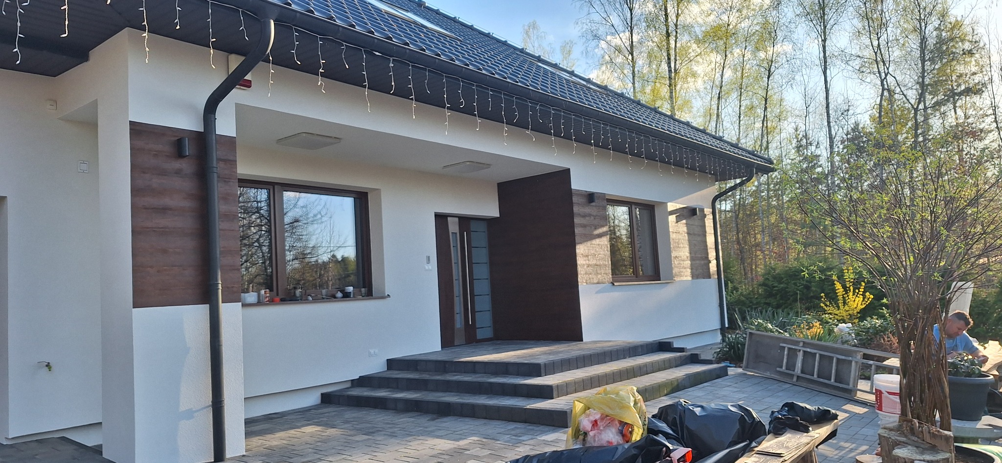 Nowoczesny dom jednorodzinny z białą elewacją i elementami dekoracyjnymi imitującymi drewno, widoczne schody wejściowe wyłożone kostką brukową, w tle drzewa i elementy ogrodu.