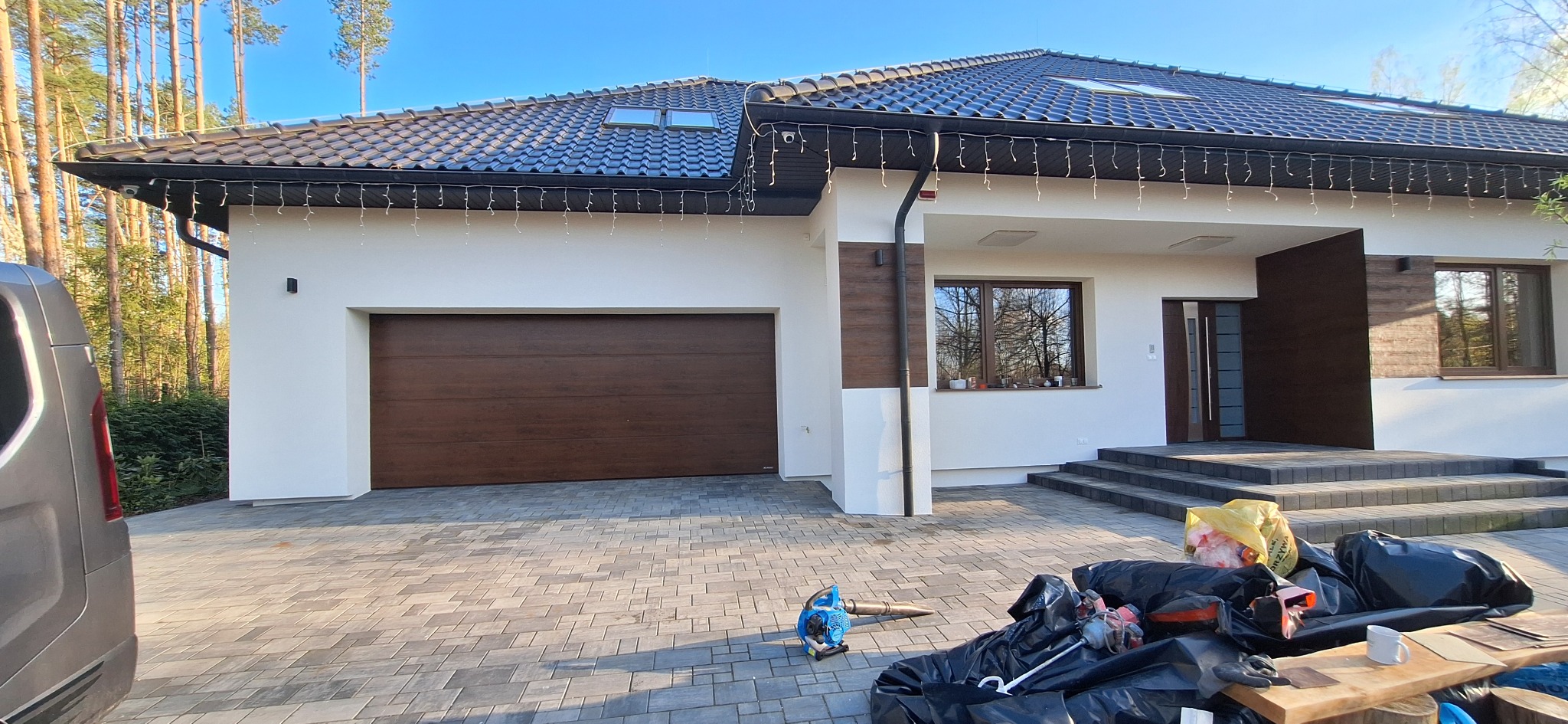 Nowoczesny dom jednorodzinny z białą elewacją, brązową bramą garażową i drzwiami wejściowymi, czarnym dachem z oknami dachowymi, oświetleniem zewnętrznym, kostką brukową na podjeździe...