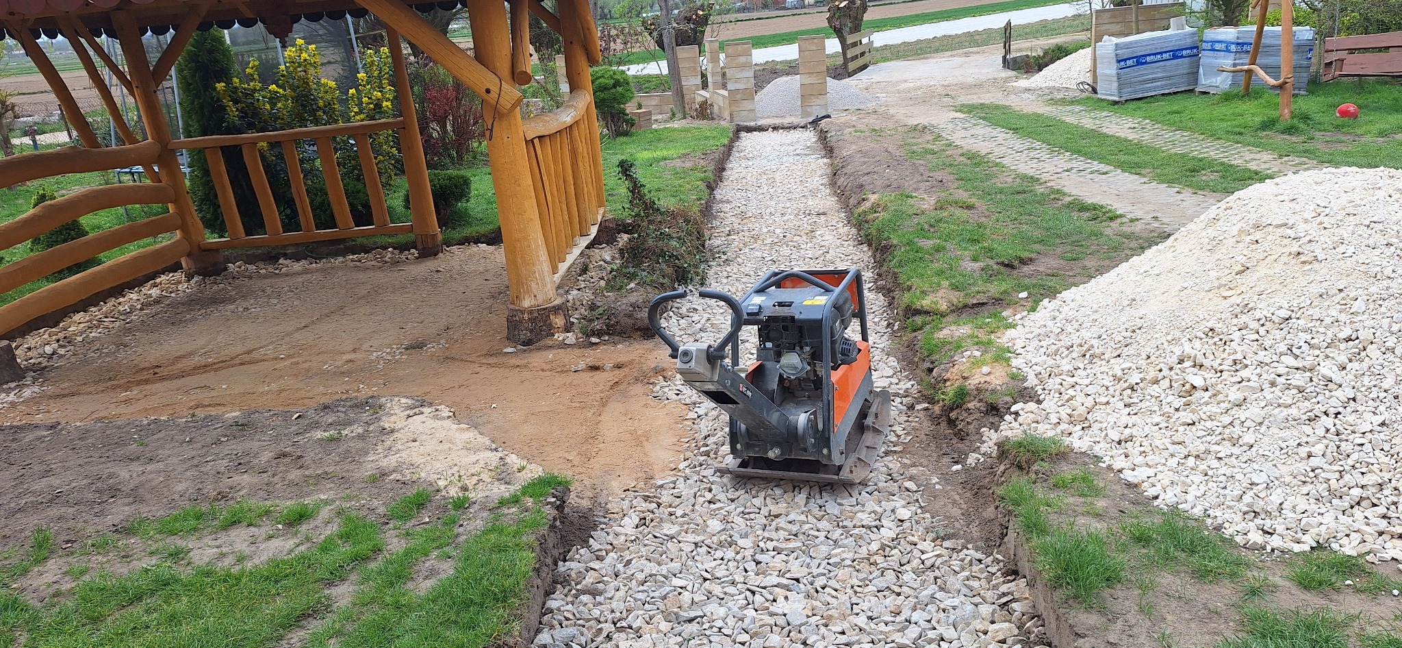 Pomarańczowa zagęszczarka wibrująca ubija warstwę białego kruszywa na przygotowanej ścieżce ogrodowej, widoczna drewniana altana z boku i stos kruszywa w tle.