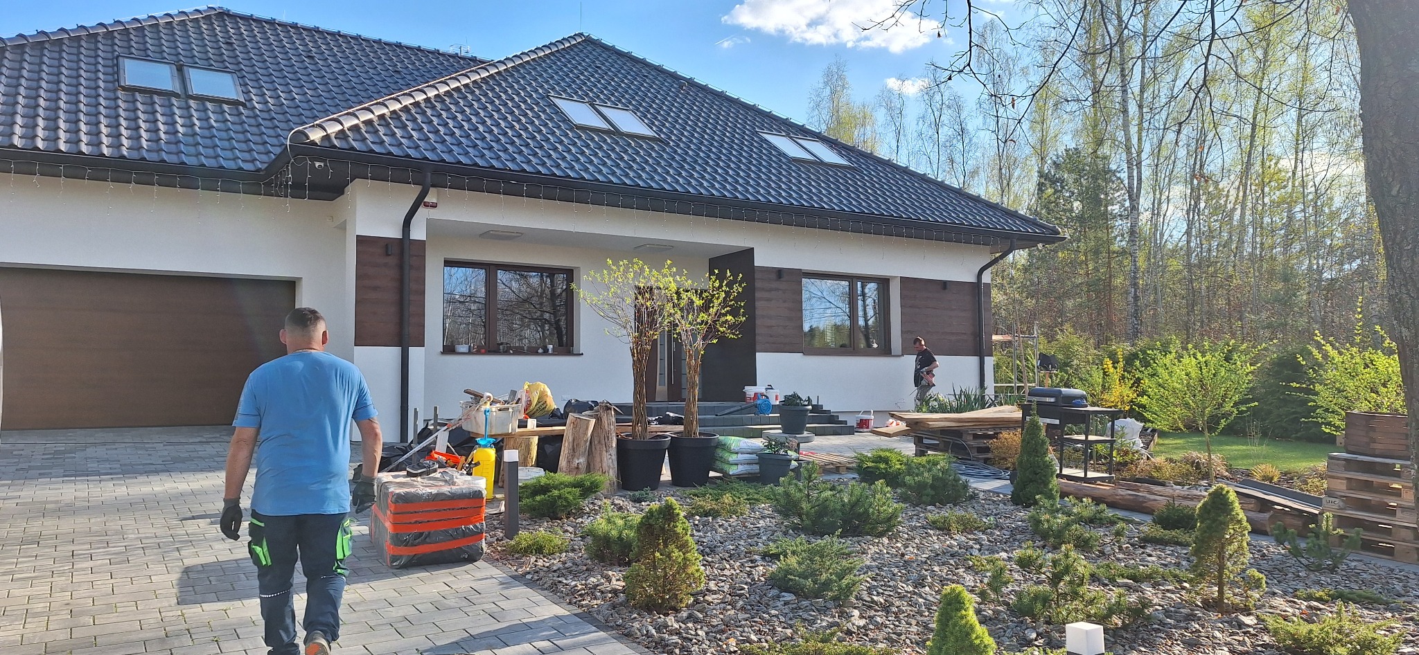 Nowoczesny dom jednorodzinny z elementami drewnianymi na elewacji, widok na ogród z kostki brukowej i roślinności, pracownik w oddali przy elewacji, drugi pracownik idzie w stronę domu.