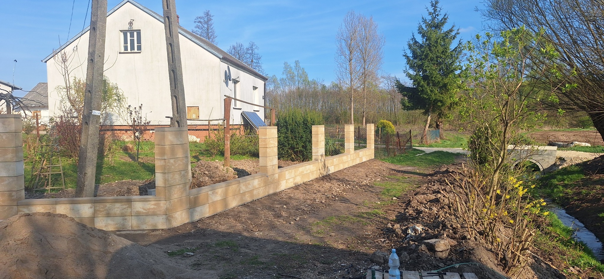 Wznoszona z beżowych bloczków podmurówka ogrodzenia na tle wiejskiego domu z instalacją solarną i zielenią, widoczne narzędzia i butelka wody na palecie.