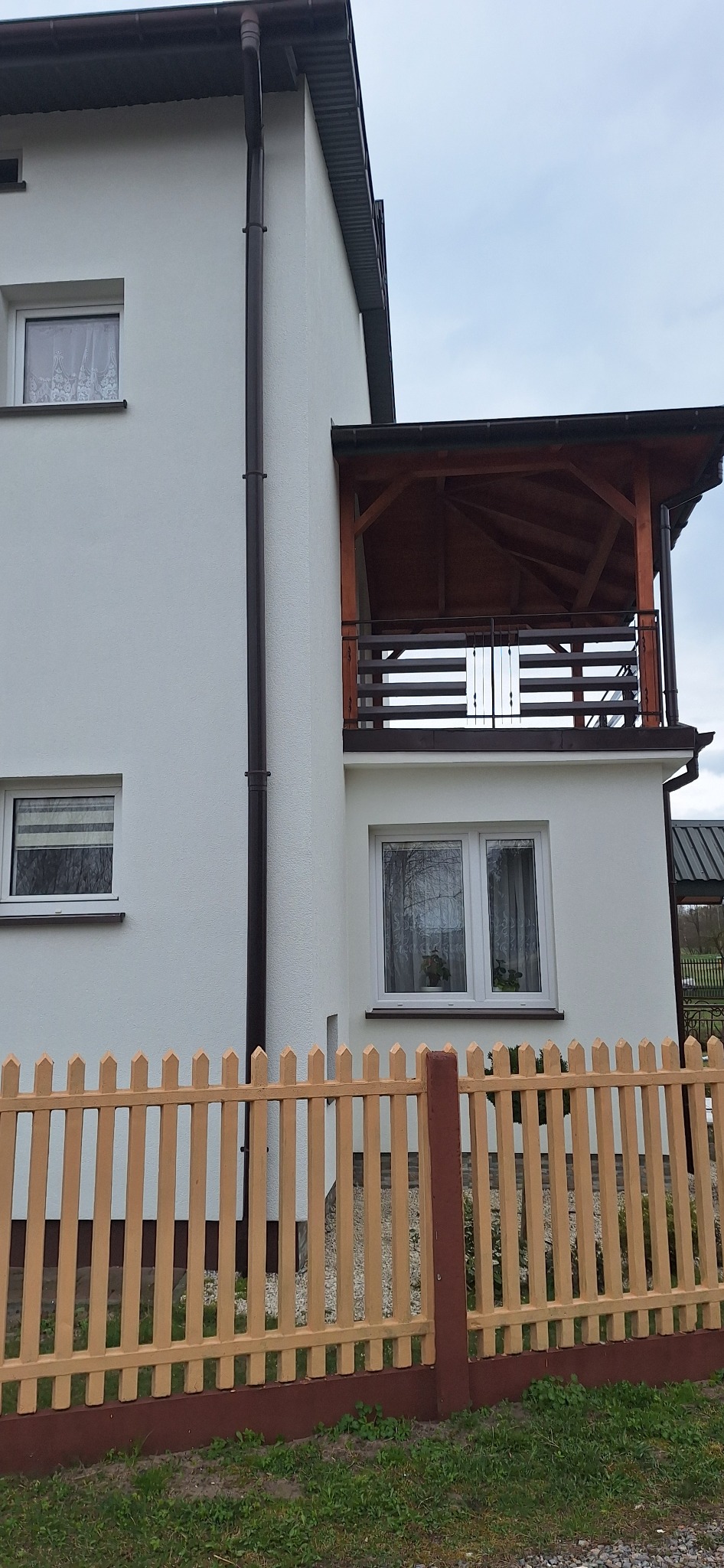 Dwukondygnacyjny dom z jasną elewacją, drewnianym balkonem z balustradą i płotem sztachetowym w odcieniach beżu i brązu.