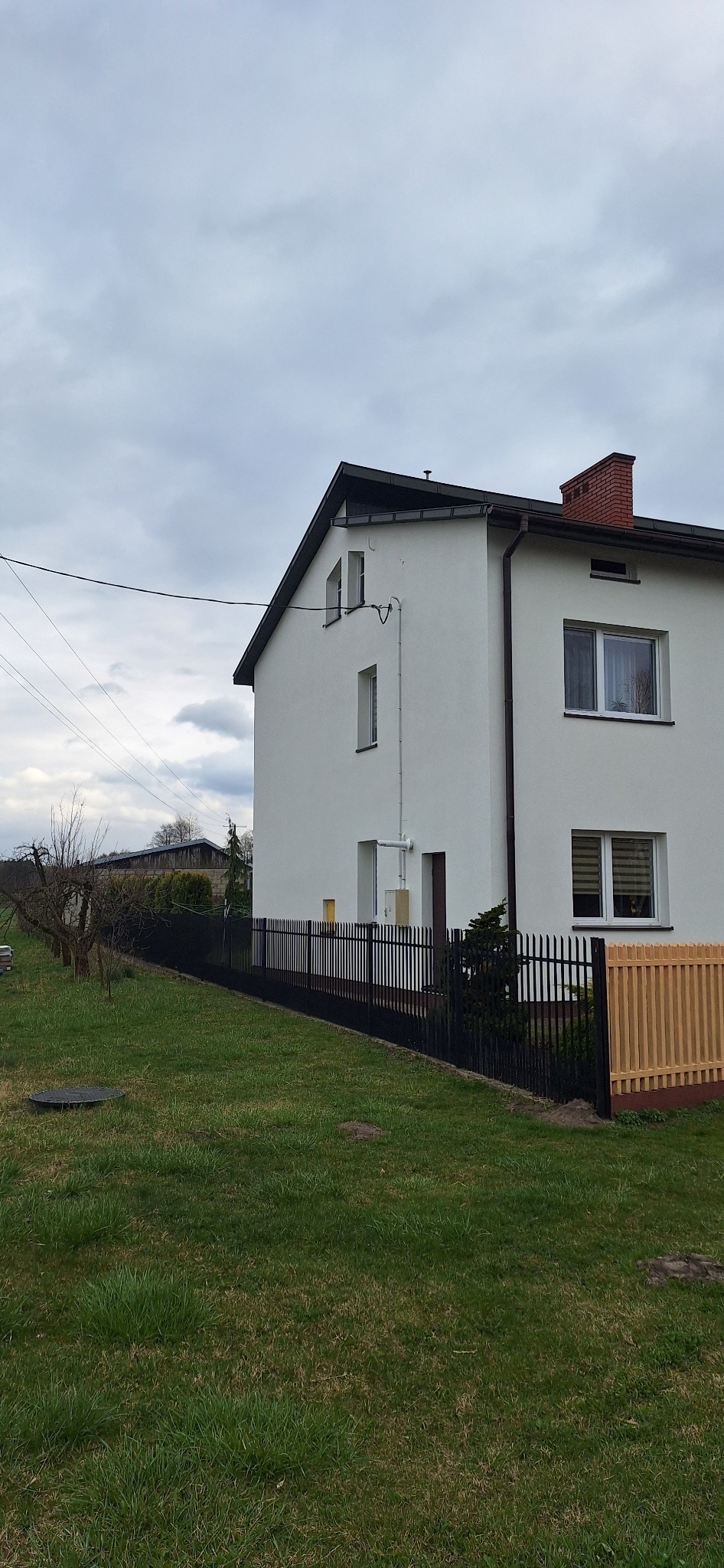 Dwukondygnacyjny dom z białą elewacją, czarną dachówką i ceglanym kominem, otoczony czarnym metalowym ogrodzeniem z drewnianym panelem, widok z poziomu trawnika w pochmurny dzień.