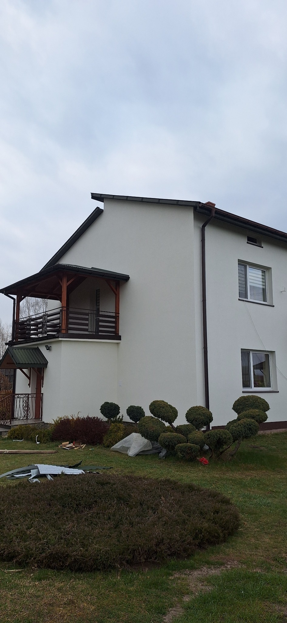 Dwukondygnacyjny dom z białymi ścianami po ociepleniu, z drewnianym balkonem i ozdobnymi krzewami w ogrodzie, widoczne pozostałości materiałów budowlanych.