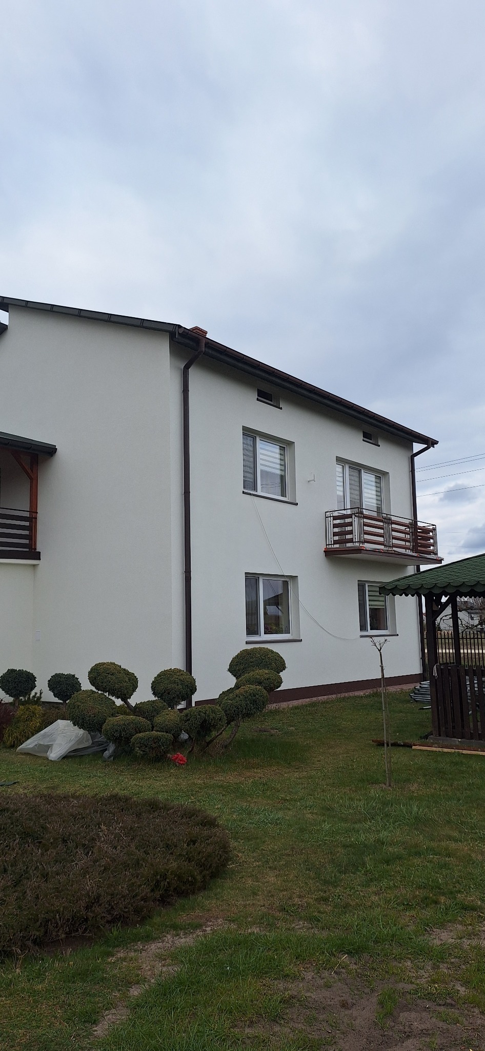 Dwupiętrowy dom z jasną elewacją, brązowymi rynnami i balkonem z drewnianymi elementami, otoczony zielonym trawnikiem z ozdobnymi krzewami w kształcie kul i małą altaną w tle.