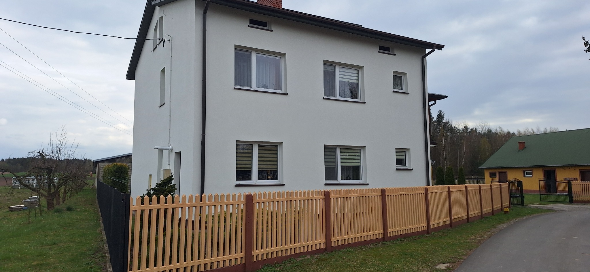 Nowoczesny, dwukondygnacyjny dom z białymi ścianami i nowym, drewnianym ogrodzeniem w kolorze jasnego drewna z ciemnobrązowymi słupkami i podmurówką, widok z ulicy.