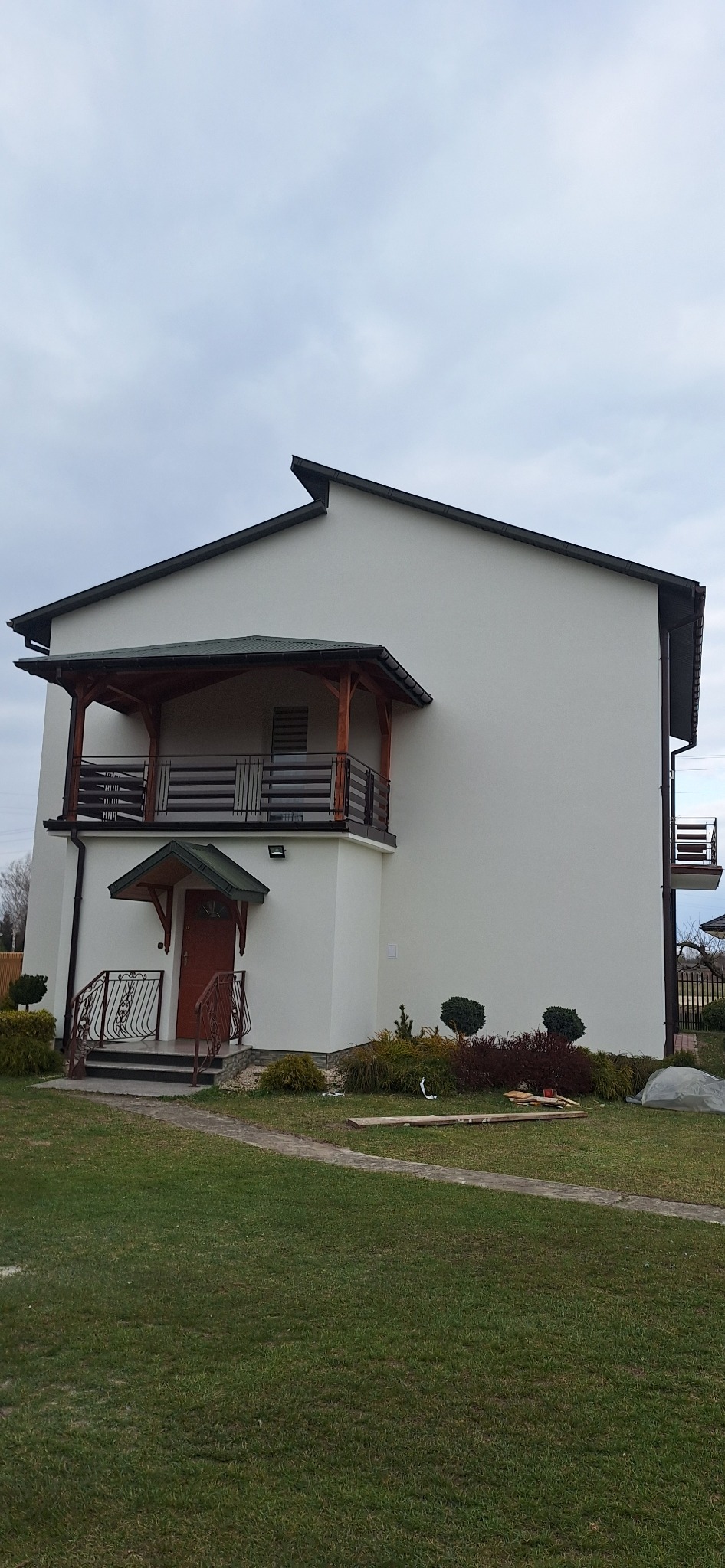Dwupiętrowy dom z białymi ścianami, drewnianymi balkonami i brązowymi balustradami, otoczony zadbanym trawnikiem z ozdobnymi krzewami, sfotografowany w pochmurny dzień.