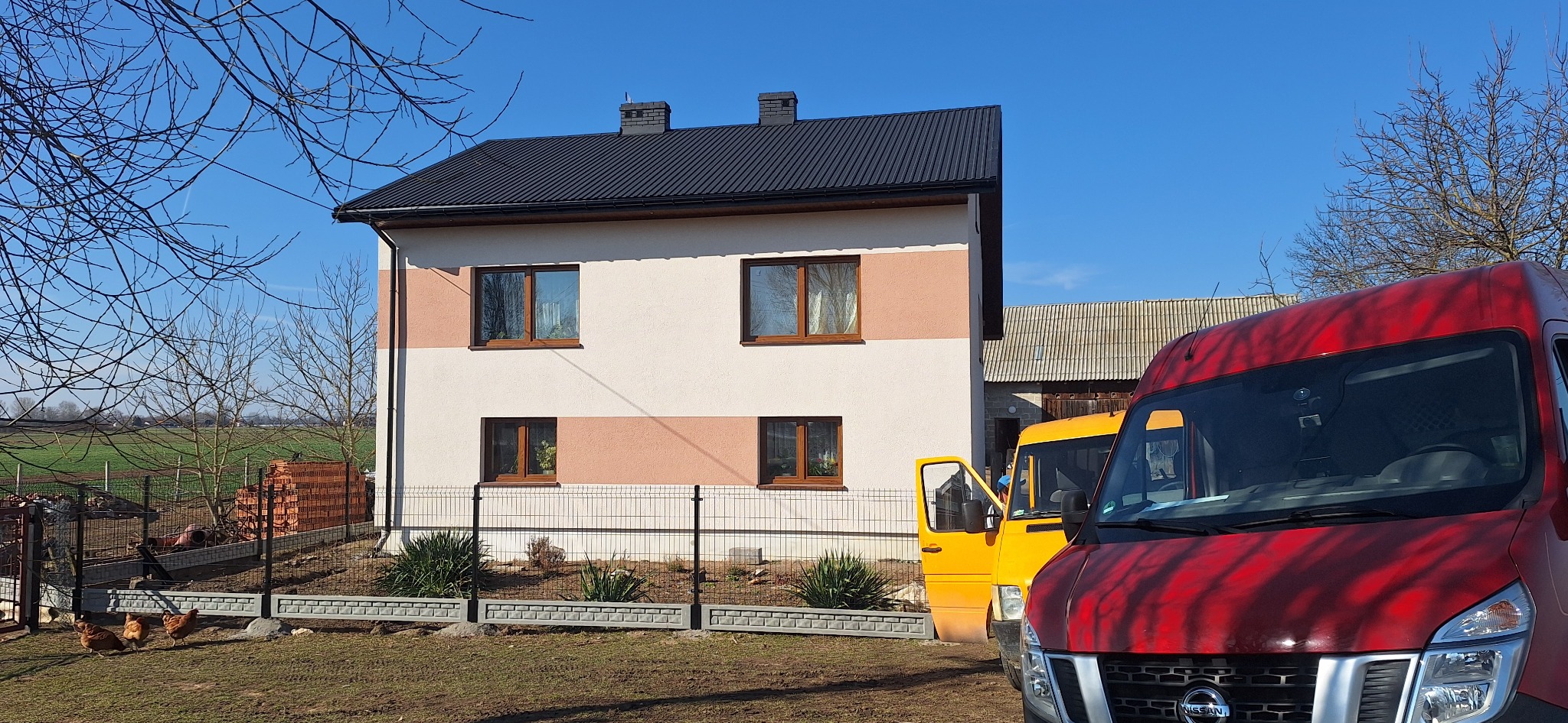 Dwukondygnacyjny dom z oknami w brązowych ramach, otoczony szarym ogrodzeniem z siatki, na podwórzu kury, w tle zaparkowane busy: czerwony Nissan i żółty Mercedes.