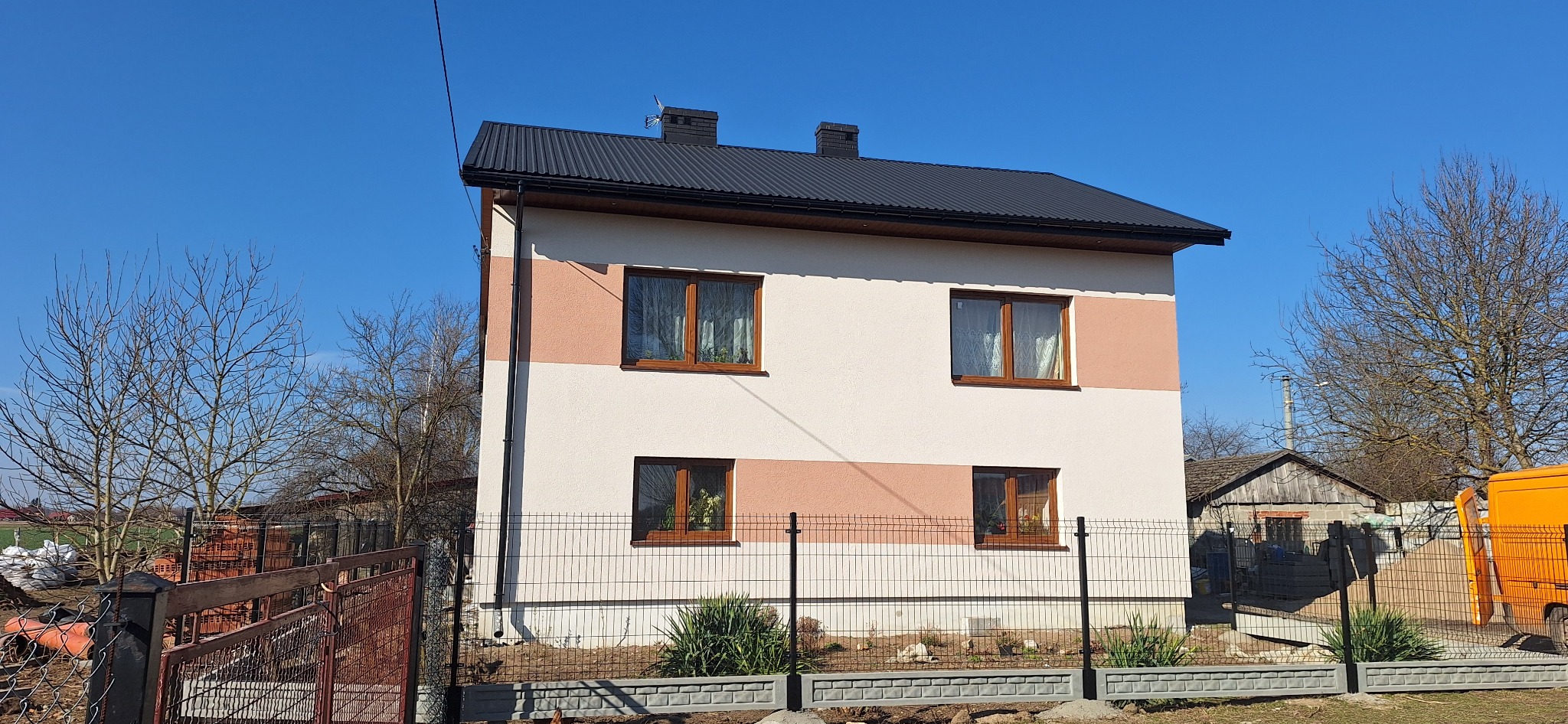 Dwukondygnacyjny dom z oknami w brązowych ramach, pomalowany w poziome pasy koloru kremowego i brzoskwiniowego, otoczony czarnym ogrodzeniem panelowym z betonową podmurówką, na tle błękitnego nieba...