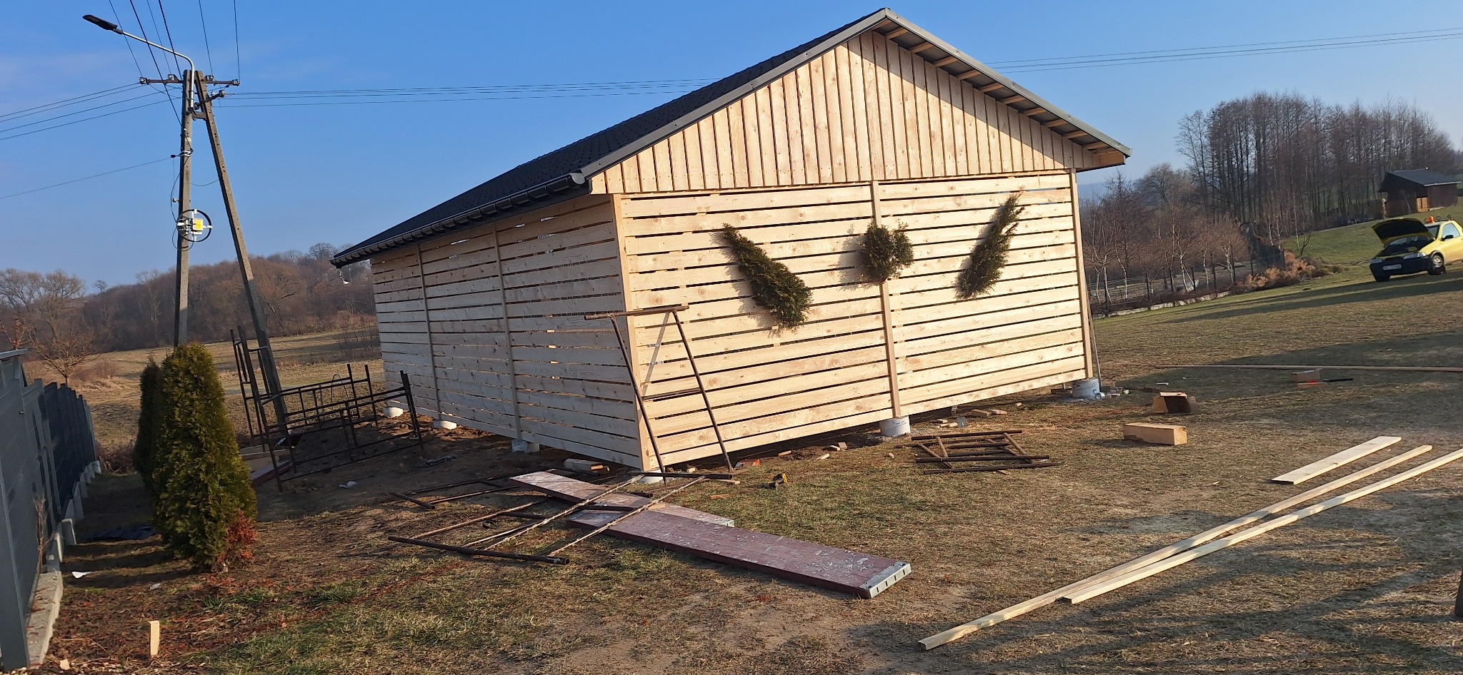 Konstrukcja szkieletowa budynku gospodarczego z drewnianą elewacją w trakcie budowy, widoczne elementy konstrukcyjne, rusztowanie, oraz otoczenie działki z drzewami i żółtym samochodem w tle.