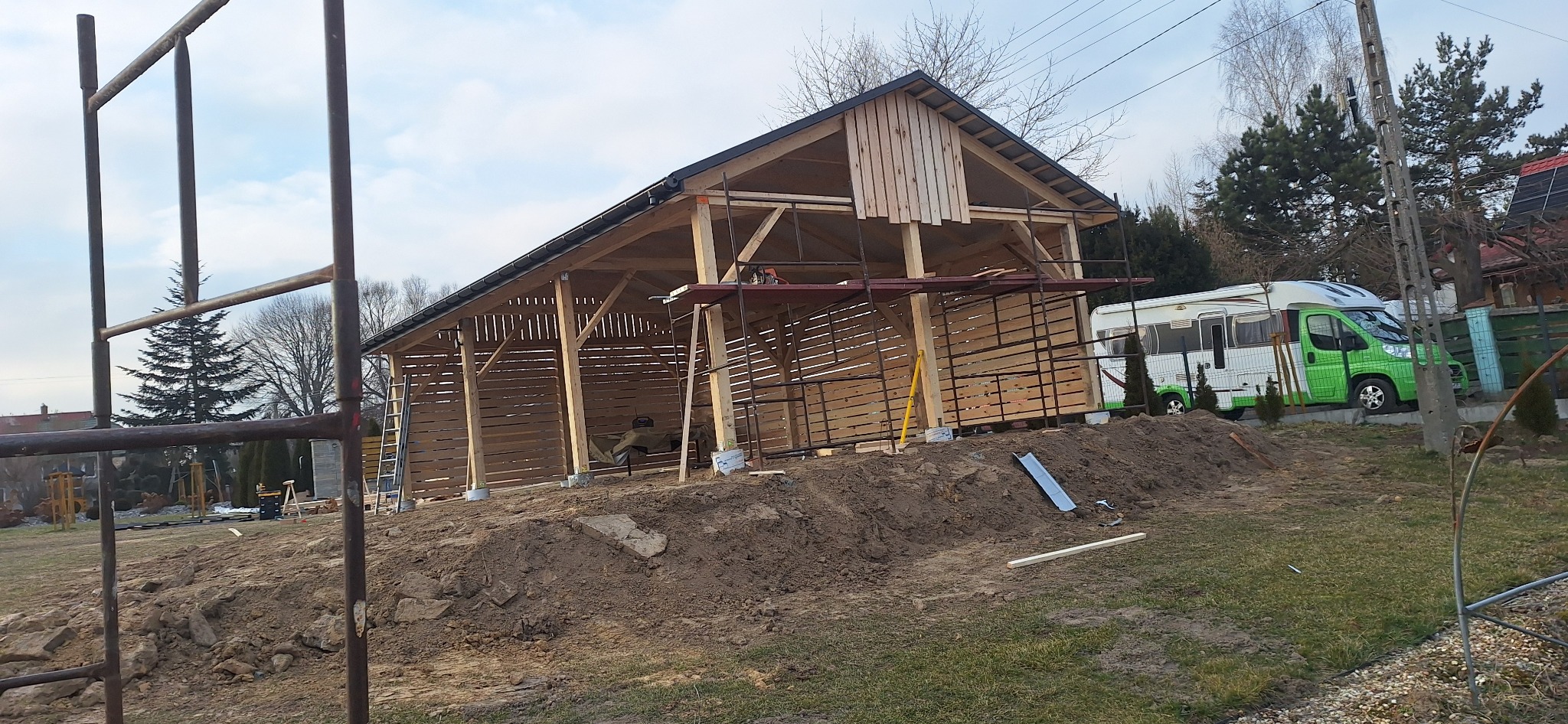 Konstrukcja drewnianego budynku gospodarczego w trakcie budowy, widoczne rusztowania i camper w tle.