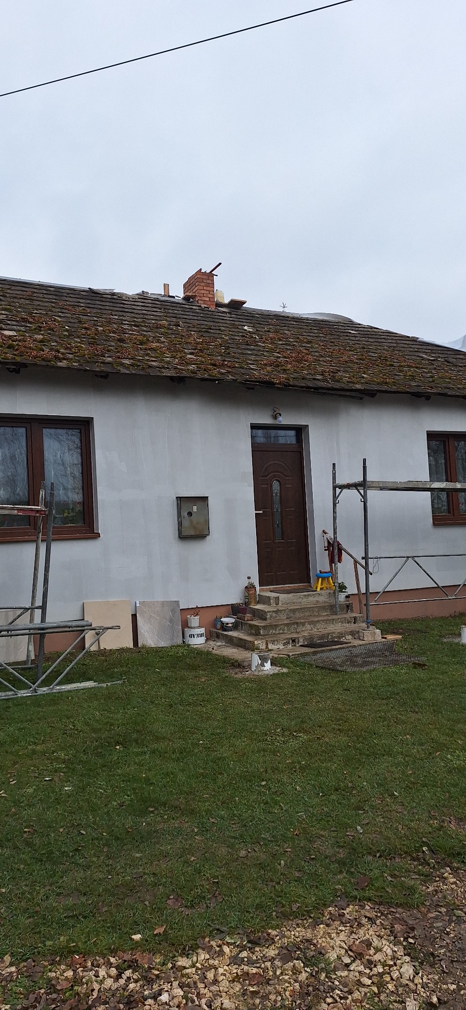 Dom jednorodzinny w trakcie prac elewacyjnych, z widocznymi rusztowaniami, przygotowanymi płytkami elewacyjnymi i świeżo pomalowaną fasadą, dach pokryty mchem