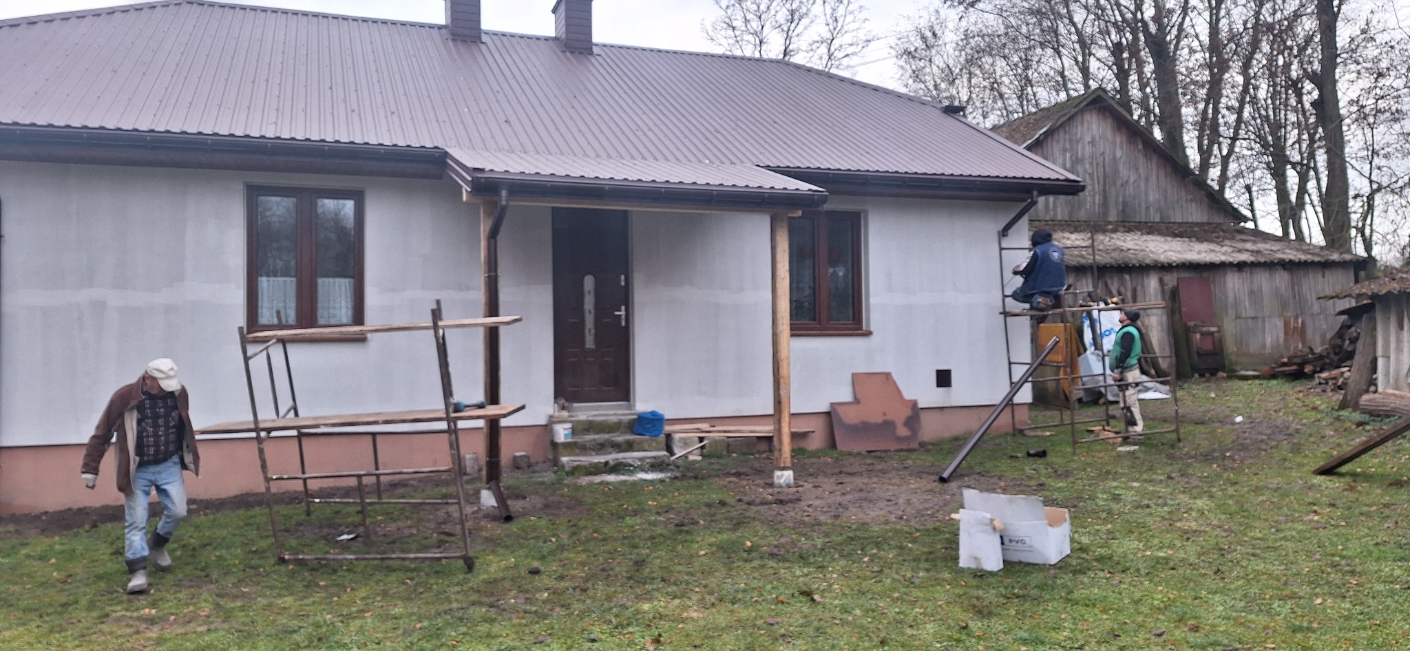 Dwóch pracowników na rusztowaniu montuje rynnę przy elewacji domu jednorodzinnego, elewacja w trakcie prac wykończeniowych, na trawniku prowizoryczne rusztowanie z desek, w tle drewniany budynek...