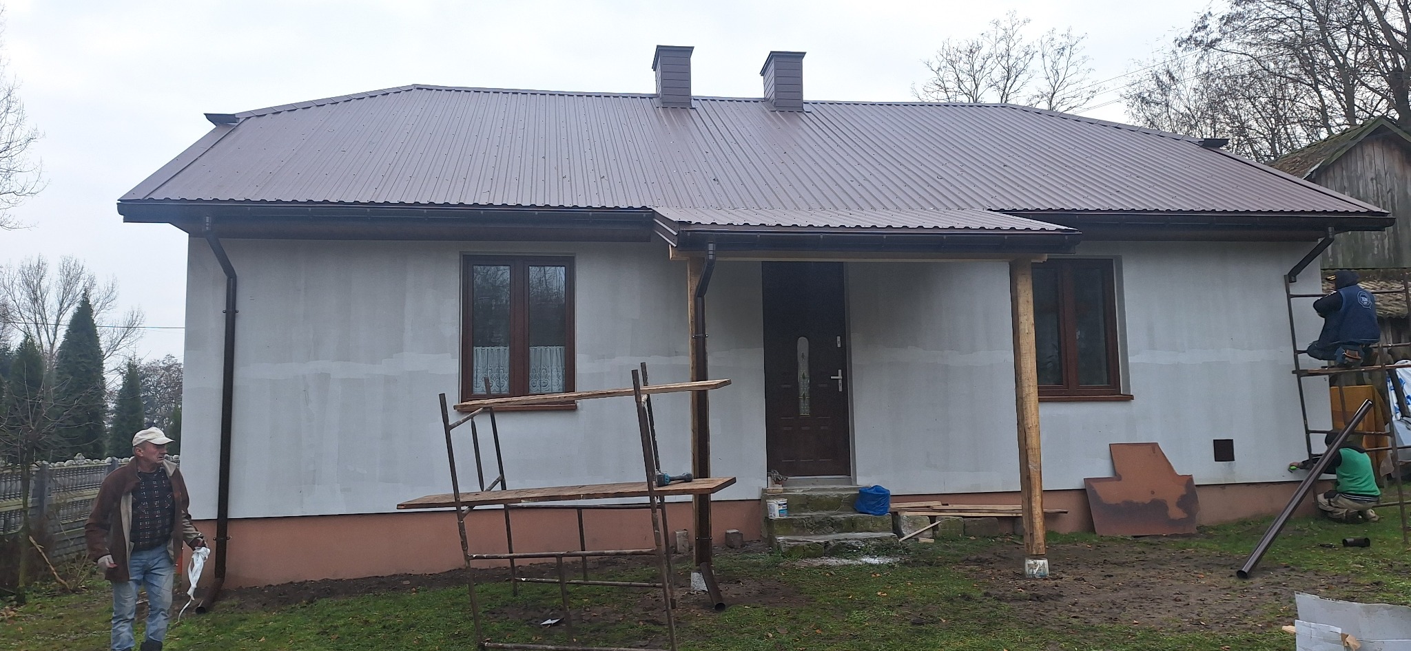 Dom jednorodzinny w trakcie prac wykończeniowych elewacji, widoczne rusztowania, pracownicy na drabinie oraz fragment ocieplonego poddasza z brązowym dachem.
