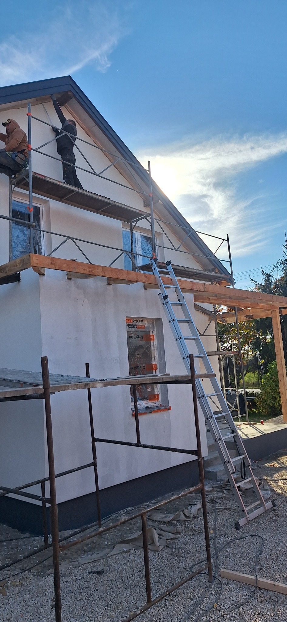 Dom jednorodzinny w trakcie prac elewacyjnych, widoczne rusztowania, drabina oparta o ścianę, okna zabezpieczone folią, dwóch pracowników na rusztowaniu.