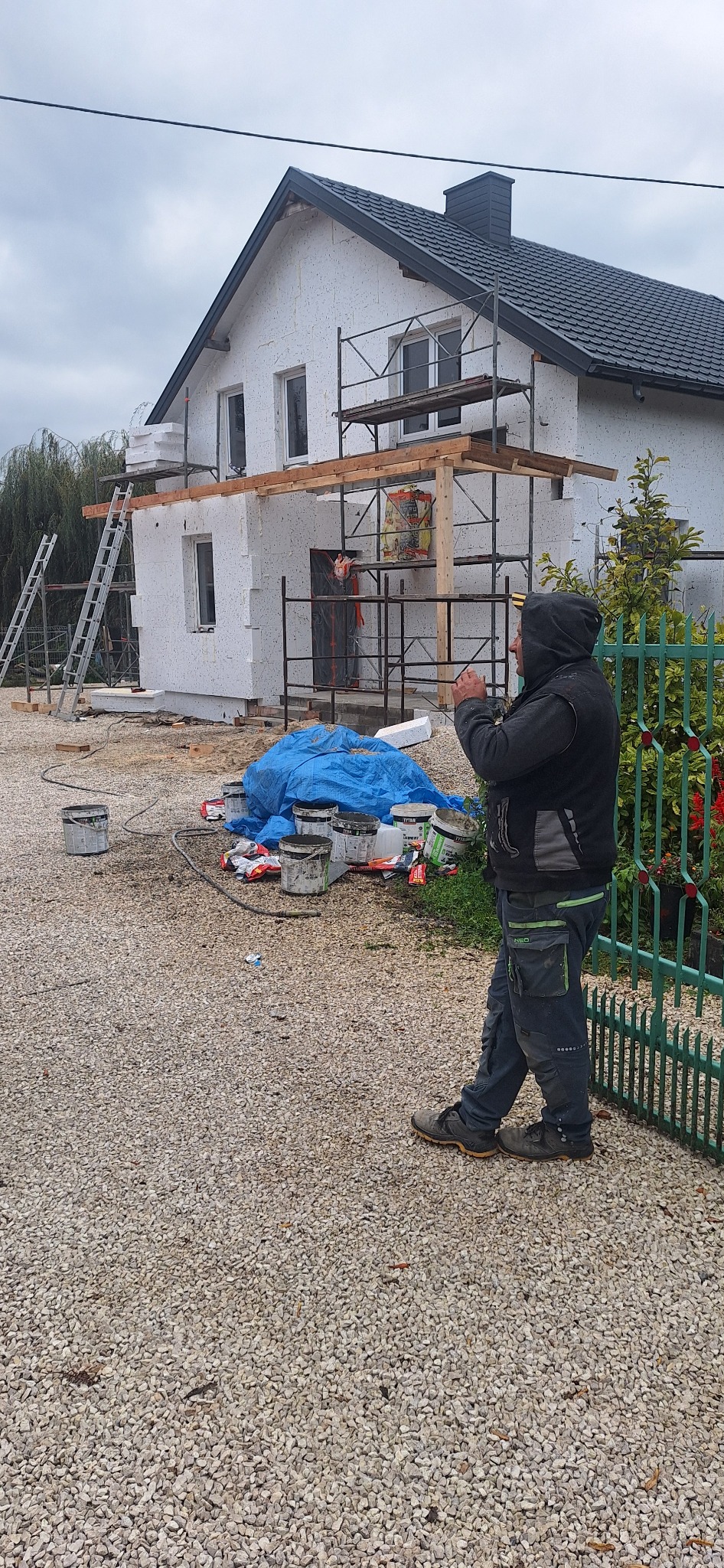 Dom jednorodzinny w trakcie prac związanych z izolacją termiczną ścian zewnętrznych, widoczne rusztowania, styropian, puszki z klejem i pracownik w odzieży roboczej